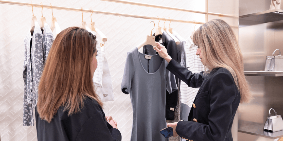 Raquel Macias, personal stylist showing a gray dress to a client inside a modern boutique, with clothing neatly displayed on wooden hangers in the background.